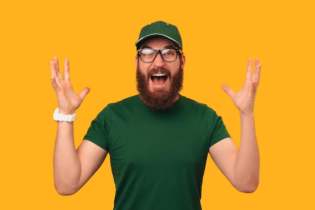 Amazed bearded man wearing green uniform is holding mouth open and arms up