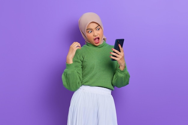 Amazed beautiful Asian woman in green sweater and hijab using a mobile phone, reading good news message isolated over purple background