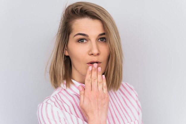 Foto stupita bella giovane donna caucasica bionda con la pelle pulita che guarda da parte con la bocca della copertura della mano che sente scioccato una buona notizia isolata sul muro bianco in studio concetto di emozione e stile di vita della gente