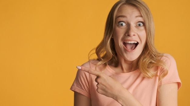 Amazed blond girl surprisingly indicates on copy space for text or promotional content over colorful background wow how exciting expression