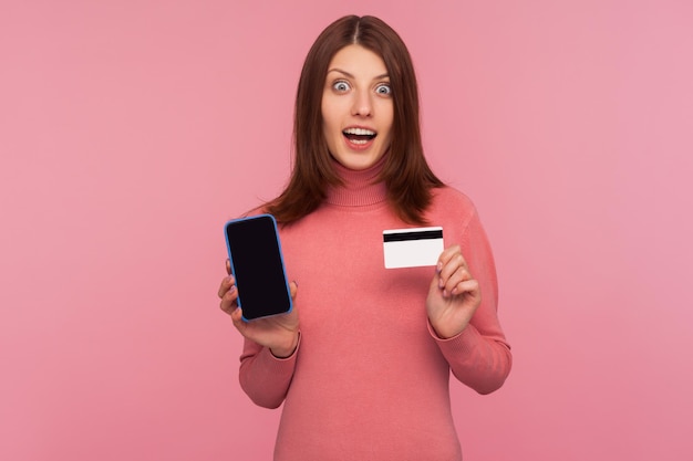 Удивленная брюнетка в розовом свитере, показывающая мобильный телефон и кредитную карту, смотрящая в камеру с потрясенным выражением лица, удивленная онлайн-банкингом. Снимок в студии на розовом фоне