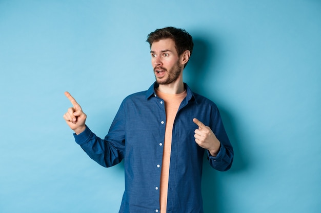 Amazed caucasian guy pointing and looking left, checking out advertisement, standing on blue background.