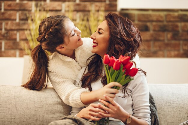 Amazed cheerful joyful charming mum is holding nice flowers and is receiving a kiss from her little cute daughter congratulates mother's day to her.