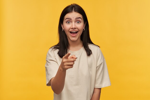 Amazed cheerful young woman with dark hair and opened mouth in white tshirt looks surprised and pointing on you over yellow wall