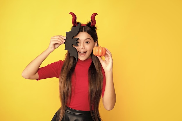 Photo amazed child in imp horns hold pumpkin and bat on yellow background cheerfulness
