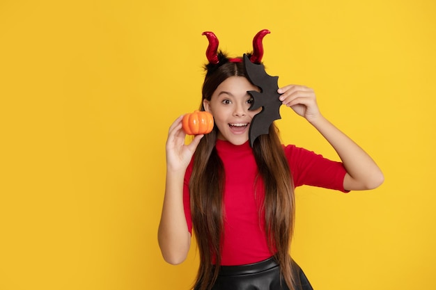 Amazed child in imp horns hold pumpkin and bat on yellow background cheerfulness