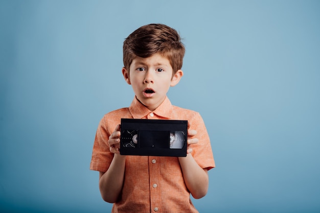 Пораженный ребенок с видеокассетой, изолированной на синем фоне, смотрит в камеру