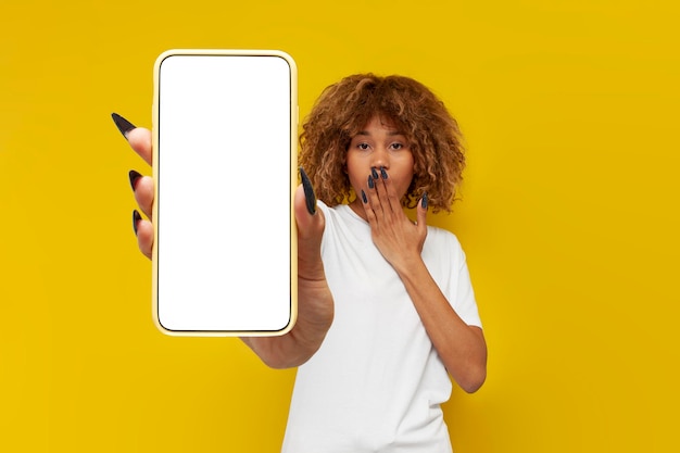 amazed curly american girl with braces shows blank smartphone screen on yellow background