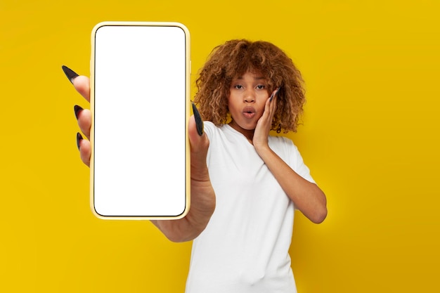 白いTシャツに中かっこをつけた驚く巻き毛のアメリカ人少女が空白のスマートフォン画面を示す