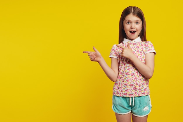 Amazed cute girl kid points away with both forefingers shows blank space