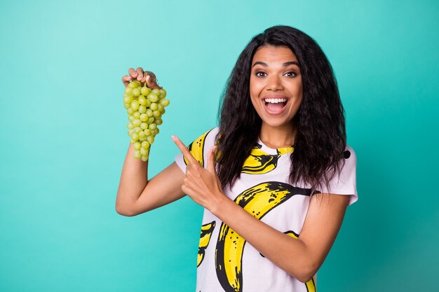 Amazed dark skin girl point finger green grapes wear pink banan print t-shirt isolated on teal color background