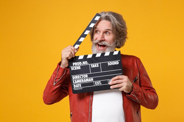 Amazed elderly gray-haired mustache bearded man in red leather jacket isolated on yellow orange background. people lifestyle concept. mock up copy space. hold classic black film making clapperboard