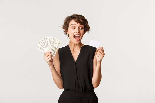 Amazed elegant woman looking at credit card, holding money, standing white.