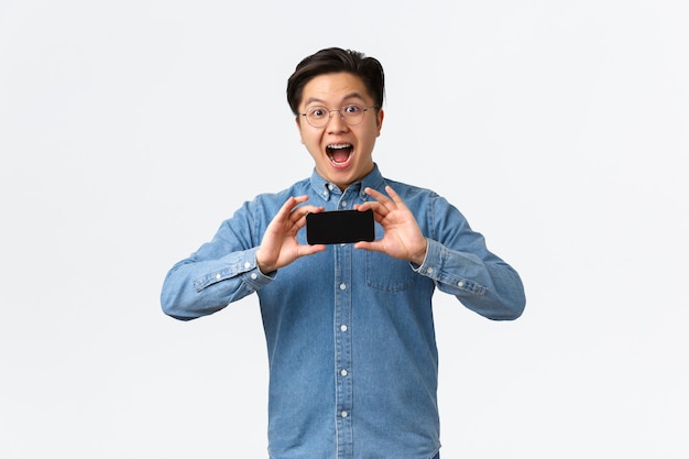 Amazed and excited asian young guy in glasses and braces showing smartphone flipped horizontally int...