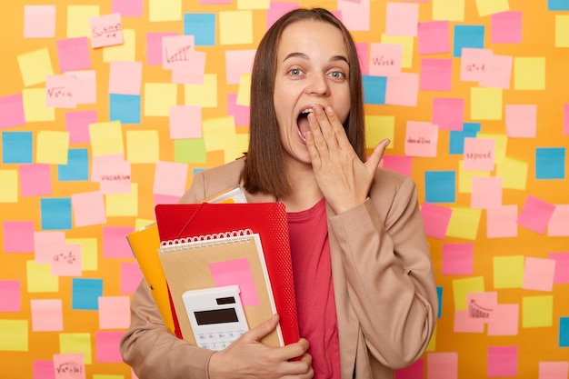 Photo amazed excited surprised woman wearing beige jacket holding paper folder being at work sees something surprised covering mouth with palm standing against yellow wall covered with stickers
