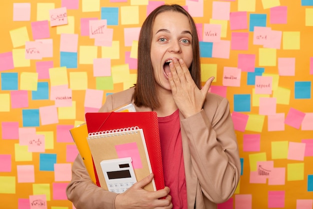 Amazed excited surprised woman wearing beige jacket holding\
paper folder being at work sees something surprised covering mouth\
with palm standing against yellow wall covered with stickers