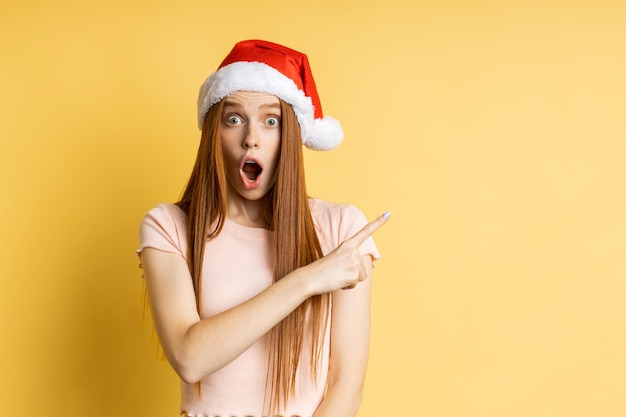 Amazed excited young redhead caucasian woman in Santa hat pointing index finger sideways, raising eyebrows and opening mouth, posing on yellow wall. Christmas, sale, advertising concept.