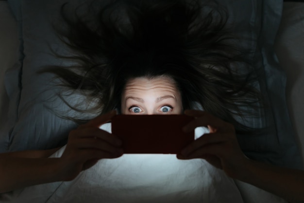 Foto volto stupito di giovane donna che fissa il suo smartphone a tarda notte a letto.