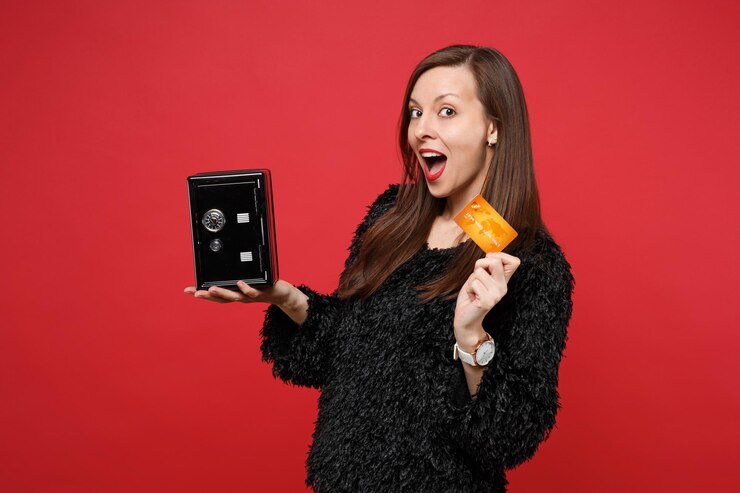 Photo amazed funny young woman in black fur sweater keeping mouth open, hold metal bank safe for money accumulation, credit bank card isolated on red background. people sincere emotions, lifestyle concept.