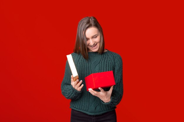 Amazed girl is opening a gift box and looking inside over red background.