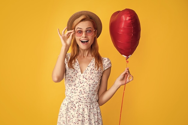 Amazed girl in straw hat and sunglasses hold love heart balloon on yellow background