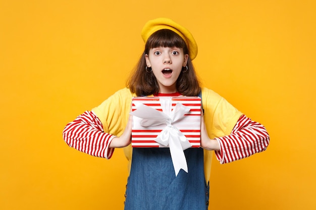 Amazed girl teenager in french beret, denim sundress with opened mouth hold red striped present box with gift ribbon isolated on yellow background. People emotions lifestyle birthday holiday concept.