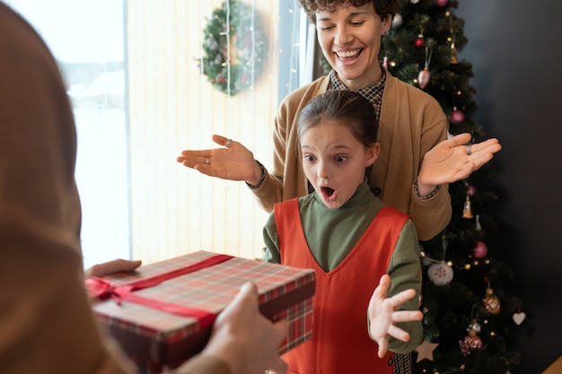 Пораженная девочка вскидывает руки и держит рот открытым, получая рождественский подарок от родителей