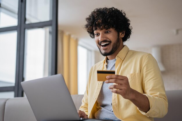 Amazed Indian man holding credit card using laptop shopping online Freelancer working from home