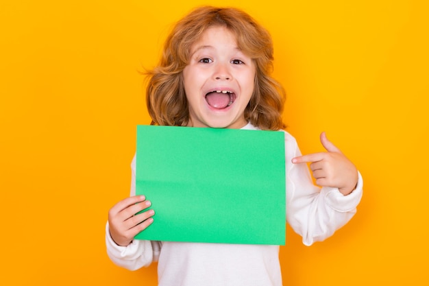 Amazed kid showing index finger on green empty sheet of paper isolated on yellow background portrait