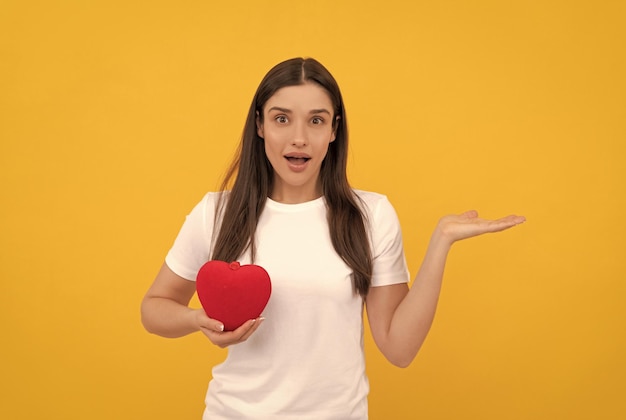 Amazed lady in white shirt with heart presenting product on yellow background copy space donation