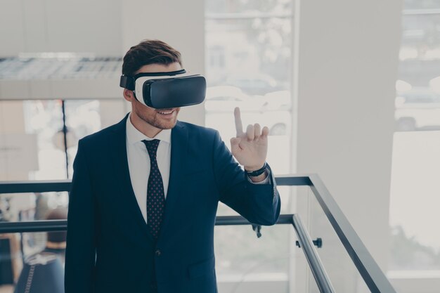 Amazed male office worker in suit being excited while trying out VR glasses, testing virtual reality for business, touching object with forefinger while exploring digital world, standing in office