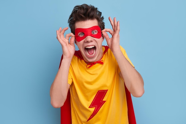 Amazed male superhero in bright mask and cloak shouting loudly and looking at camera on blue background in studio