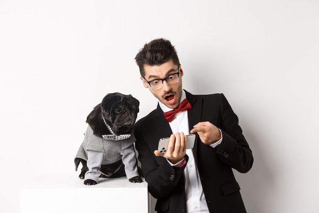 Amazed man showing video to dog, pointing at mobile phone and look excited while pug sitting unbothered in costume, white background.