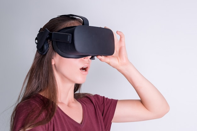 Amazed positive woman wearing virtual reality goggles headset, vr box. Connection, technology, new generation, progress concept. Girl surprised by something in virtual reality. Studio shot on gray