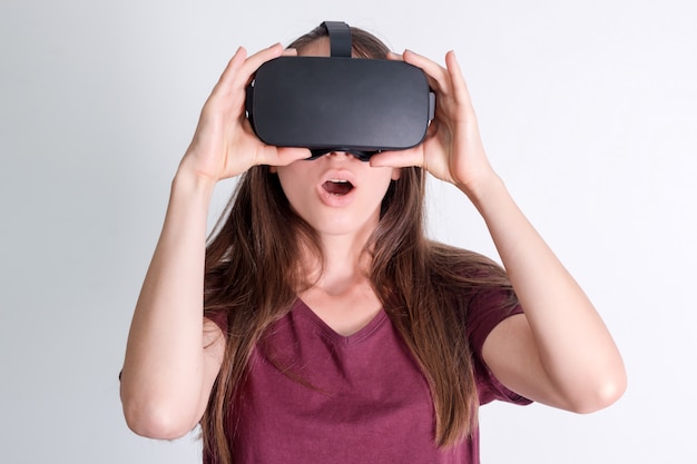 Amazed positive woman wearing virtual reality goggles headset, vr box. Connection, technology, new generation, progress concept. Girl surprised by something in virtual reality. Studio shot on gray