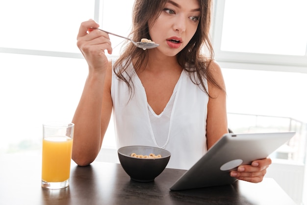 Amazed pretty young woman having breakfast and using tablet on the kitchen