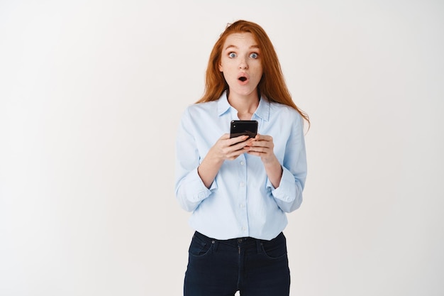 Amazed redhead girl reading promo offer online, holding smartphone and looking surprised at front, white wall