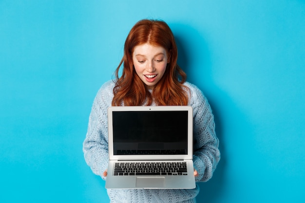 Пораженная рыжая девушка смотрит на экран ноутбука и выглядит впечатленным, показывая дисплей компьютера, стоя на синем фоне.