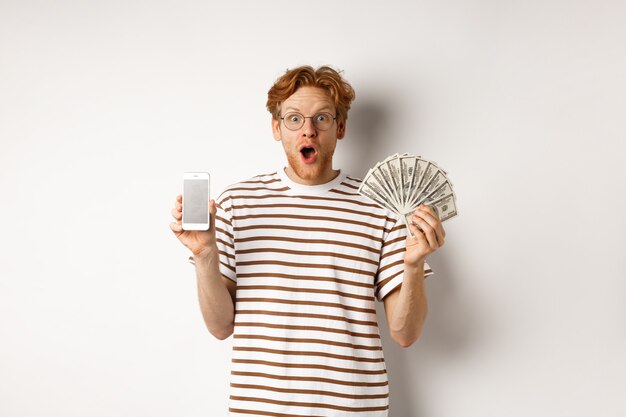 Amazed redhead man showing smartphone app on blank screen and money, winning prize cash online, standing over white background