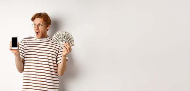 Фото Изумленный рыжеволосый мужчина показывает приложение для смартфона на пустом экране и выигрывает денежные призы онлайн