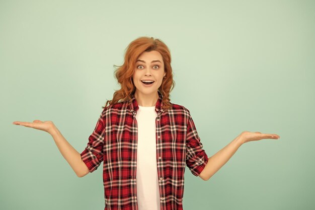 Amazed redhead woman in checkered shirt presenting product on blue background