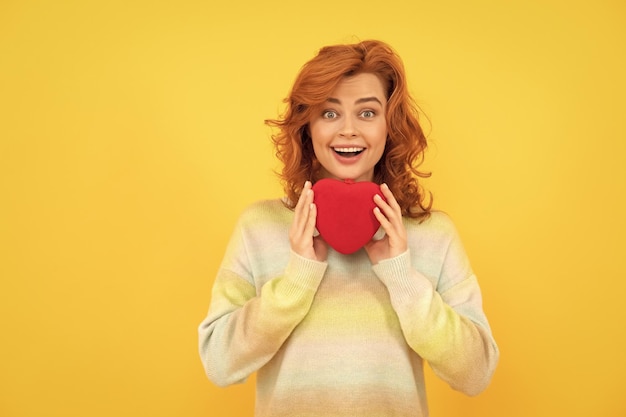 Amazed redhead woman with red heart on yellow background love