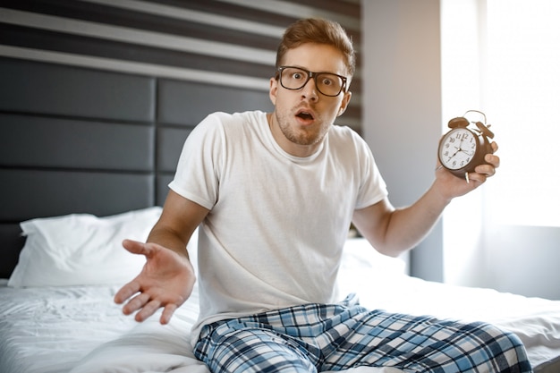 Photo amazed scared young man on bed this morning. he look on camera and hold clock. he overslept.