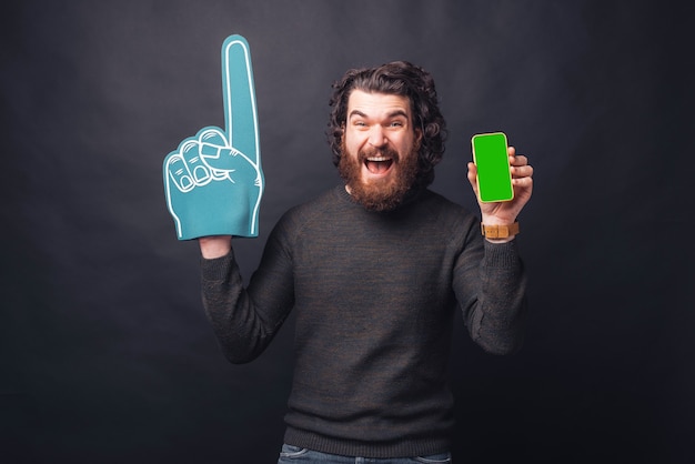 Amazed screaming young bearded man with fan glove showing green screen on phone