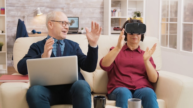 Photo amazed senior woman while using virtual reality goggles on sofa with her husband next to her using laptop.