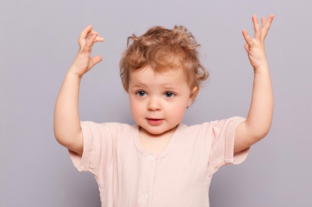 Amazed serious little caucasian toddler girl with blond wavy hair wearing casual shirt standing with raised arms showing how high she will grow posing isolated over gray background