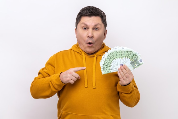 Amazed shocked man holding pointing at many euro banknotes looking at camera with open mouth