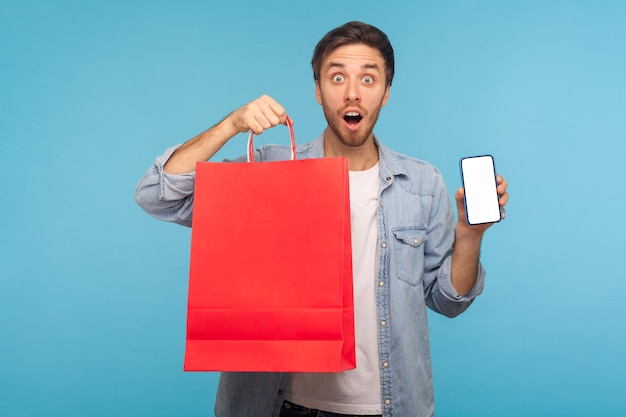 Amazed shocked man holding shopping bags and cell phone with mock up blank display for advertisement of online store mobile app goods purchase and delivery studio shot isolated on blue background