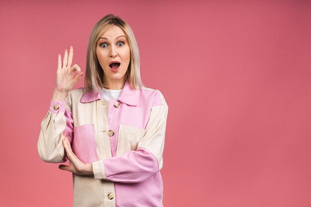 Amazed shocked surprised young blonde woman looking at camera isolated on pink background. Ok sign.