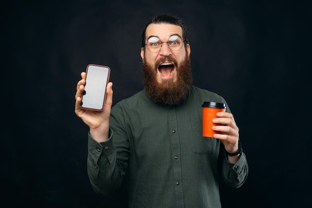 Amazed and shocked young man with beard showing smartphone screen and hodling cup of coffee take away